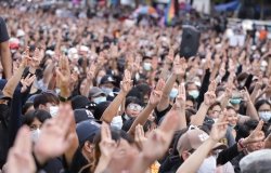 A crowd of people gathered at a protest, lifting their hands with three fingers extended in a gesture of protest against the government.