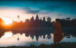 A monk stands in the foreground looking out at a temple at sunrise.