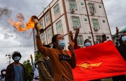 A group of protestors march with the flag of the National League for Democracy, one of them is carrying a lit torch.