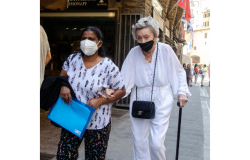 two people walking, both wearing masks, one holding a cane, the other holding a folder
