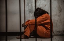 Woman in prison wearing orange jumpsuit sitting behind bars.