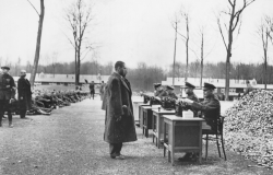 Gestapo officials recording data on incoming prisoners at a German concentration camp. Many others are seated on the ground