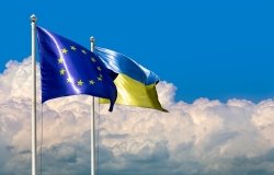 Two flags of Ukraine and the European Union on flagpoles at blue cloudy sky.